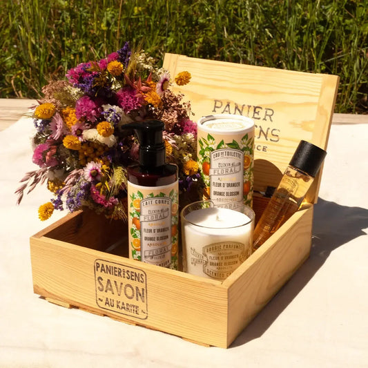 Luxury organic skincare gift set with floral bouquet, candles, and body products in wooden box outdoors