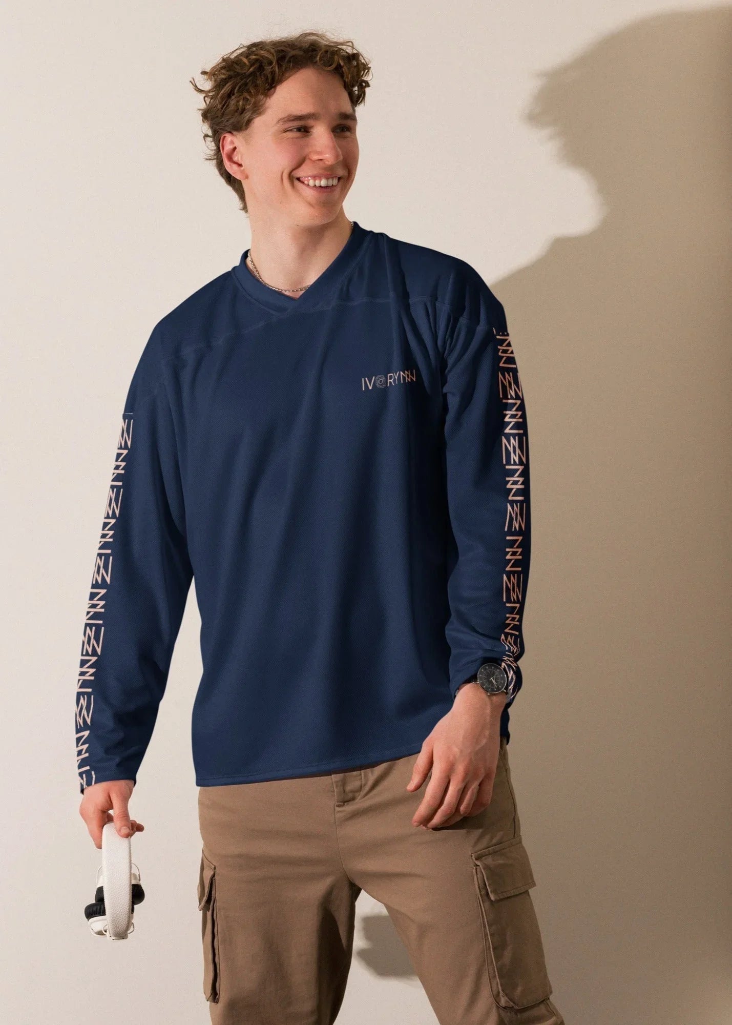 Smiling man wearing IVORYNN navy blue long sleeve sportswear top with logo, khaki pants, holding headphones