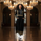 Woman in a black and gold long dress with hood, walking under crystal chandeliers in a grand hall
