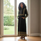 Woman in black and gold patterned dress stands barefoot by large window with garden view